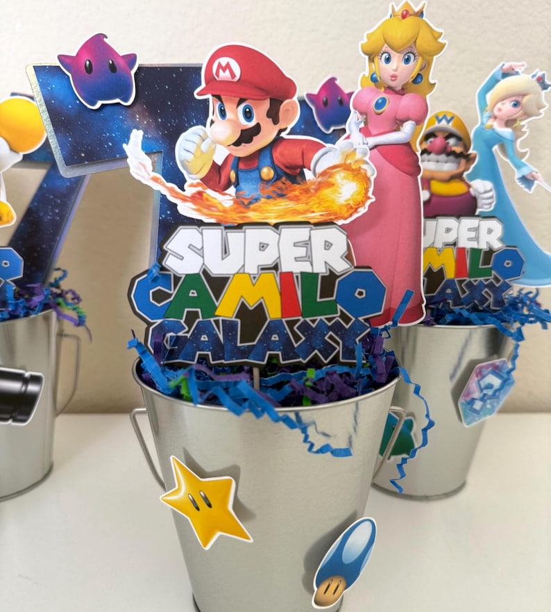 May include: A silver metal bucket centrepiece featuring a cutout of Mario and Princess Peach with the text Super Camilo Galaxy. The bucket is filled with blue and purple paper shreds and decorated with small star and mushroom character stickers.