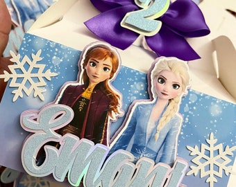 May include: A white cardboard box decorated with a purple bow, a blue number 2, and images of Elsa and Anna from the movie Frozen. The box has a snowflake design and the name "Emma" written in blue glitter letters.
