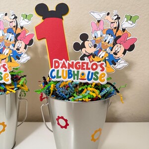 May include: Two silver metal buckets decorated with colourful gear stickers. Each bucket is filled with shredded paper and topped with a Mickey Mouse-themed centrepiece. The centrepiece features a large red number 1 and the text "D'Angelo's Clubhouse".
