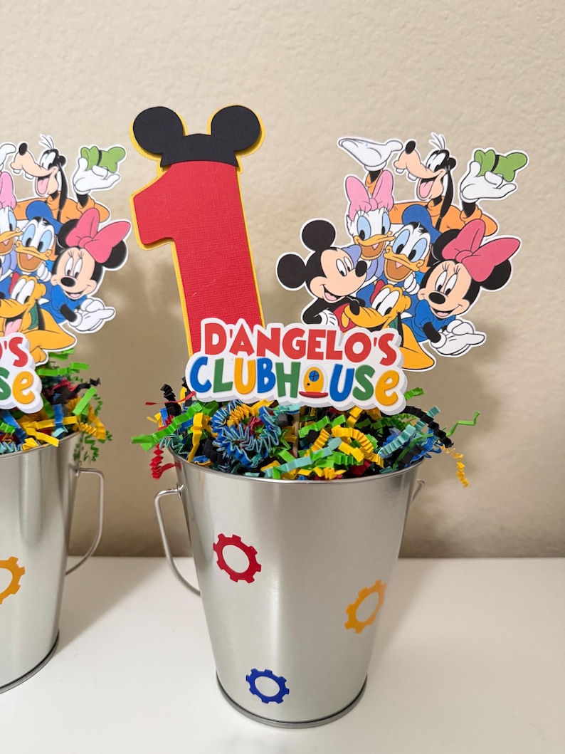 May include: Two silver metal buckets decorated with colourful gear stickers. Each bucket is filled with shredded paper and topped with a Mickey Mouse-themed centrepiece. The centrepiece features a large red number 1 and the text "D'Angelo's Clubhouse".