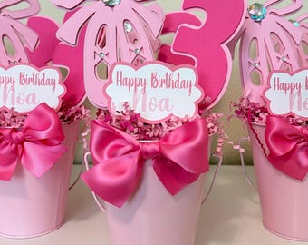 Op de afbeelding: Roze metalen emmers met decoraties voor een verjaardag. Elke emmer heeft een grote roze satijnen strik, een papieren uitsnede van een balletschoen en een bord met de tekst "Happy Birthday Noa". Een groot cijfer "3" is ook aanwezig.