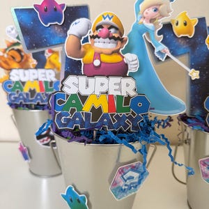 May include: A silver metal bucket centrepiece featuring a cutout of Wario and Rosalina from Super Mario Galaxy. The topper displays the text SUPER CAMILO GALAXY in colourful block letters. Blue paper shred fills the bucket, accented with star cutouts.