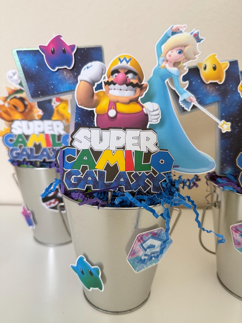 May include: A silver metal bucket centrepiece featuring a cutout of Wario and Rosalina from Super Mario Galaxy. The topper displays the text SUPER CAMILO GALAXY in colourful block letters. Blue paper shred fills the bucket, accented with star cutouts.