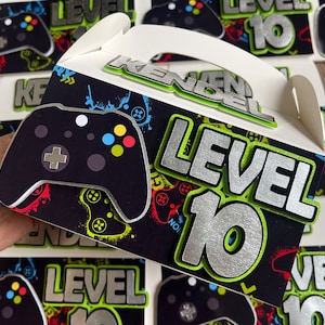 May include: A black and green party favor box with a silver and green "Level 10" design. The box has a handle and a black and white video game controller graphic.