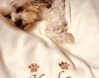 May include: A white plush dog blanket with brown paw prints and the name "Koda" embroidered on it. A small dog is sleeping on the blanket.