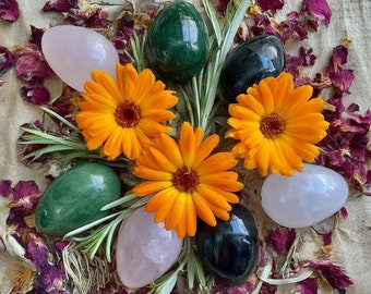 May include: A collection of seven polished stones, including green, black, and pink, arranged on a fabric background with orange flowers. The stones are smooth and have a natural, organic look.