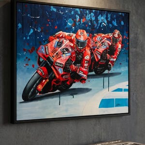 May include: A framed artwork showing two red Ducati motorbikes in a race. The riders are in red suits and helmets. The background features blue and white paint splashes. The artwork is presented in a black frame.