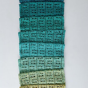 Op de afbeelding: Een gehaakte sjaal in tinten blauw, groen en geel. De sjaal is gemaakt van een delicaat, kantpatroon en is perfect om een ​​kleuraccent toe te voegen aan elke outfit.