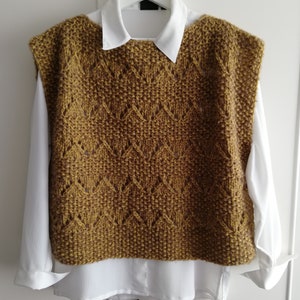 May include: A sleeveless knitted vest in a mustard yellow color with a diamond pattern. The vest has a wide neckline and is made of a soft, textured yarn.