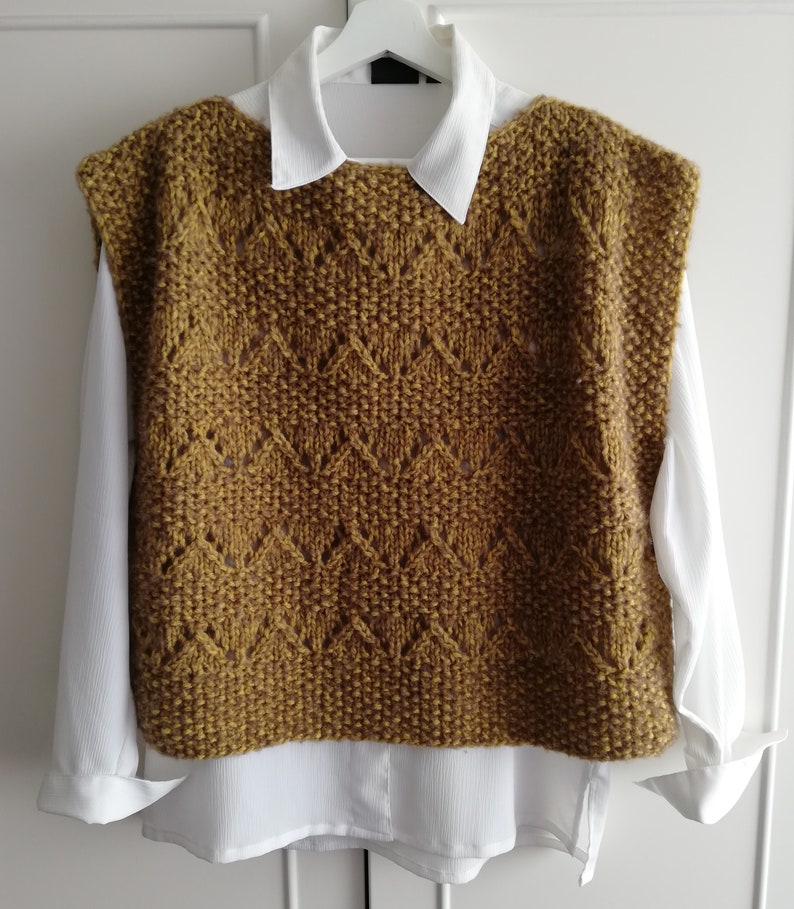 May include: A sleeveless knitted vest in a mustard yellow color with a diamond pattern. The vest has a wide neckline and is made of a soft, textured yarn.