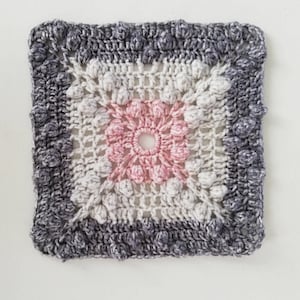 May include: A crocheted square with a pink and white center and a gray border. The square is made with a textured stitch that creates a raised effect.