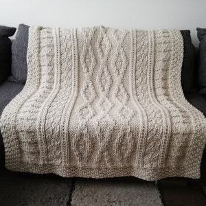 Puede incluir: Una manta de punto de color crema con un patrón de punto de cable. La manta está colocada sobre un sofá gris.