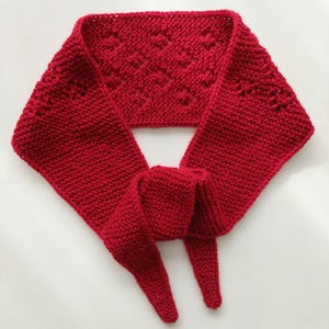 Könnte beinhalten: Ein leuchtend roter Strickschal mit strukturiertem Muster. Der Schal ist aus weichem Garn gefertigt und hat ein Rautenmuster in der Mitte. Er ist zum Binden um den Hals konzipiert und hat lange, spitz zulaufende Enden. Ein gemütliches Accessoire für kalte Tage.