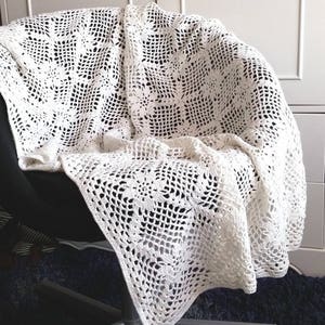 Op de afbeelding: Een witte, gehaakte deken gedrapeerd over een zwarte stoel. De deken heeft een herhalend bloemmotief binnen een vierkant raster. Het open ontwerp creëert een delicate, luchtige textuur. De randen van de deken zijn afgewerkt met een eenvoudige rand.