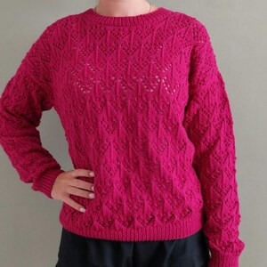 Op de afbeelding: Een levendige fuchsia trui met lange mouwen en een ingewikkeld, herhalend ruitvormig kabelbreipatroon. Het kledingstuk heeft een klassieke ronde hals, geribbelde boorden aan de mouwen en een geribbelde zoom met een textuurrijk opengewerkt ontwerp.