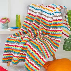 Op de afbeelding: Een gehaakte deken met een herhalend chevronpatroon in levendige horizontale strepen van wit, beige, geel, oranje, teal en magenta. Het textiel heeft een gestructureerde, golvende steek en is gedrapeerd over een stoel.