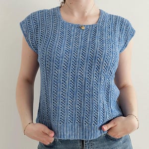Op de afbeelding: Een lichtblauw gebreid topje met een verticaal chevron-steekpatroon, korte mouwen en een ronde halslijn. Het kledingstuk is gemaakt van getextureerd garen met subtiele variaties in blauwtinten en heeft een geribbelde zoom.
