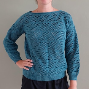 Op de afbeelding: Een teal-kleurige trui met lange mouwen en een ingewikkeld opengewerkt breipatroon met een herhalend chevronontwerp. Het kledingstuk heeft een brede boothals en geribbelde boorden bij de polsen en de zoom, gemaakt van effen blauwgroen garen.