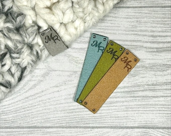 May include: A close-up of several rectangular tags with the initials "MR" embossed on them. The tags are in various colors, including gray, blue, green, and tan. The tags are placed near a white and gray knitted item.