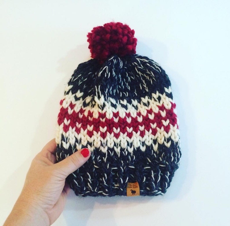 May include: A knitted beanie in navy blue, white, and red with a burgundy pom-pom. The hat features a textured pattern and a small leather tag. The hat is held by a person.
