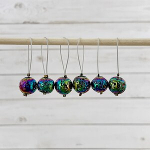 May include: Six iridescent, textured glass beads with a rainbow sheen are suspended from a wooden dowel. Each bead is round and has a small loop at the top, suggesting they are decorative or for crafting.
