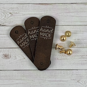 May include: Three brown leather tags with the text "hand made" in white lettering. The tags are stacked on top of each other and have a hole punched in the top. There are four gold metal rivets on a white wooden surface.