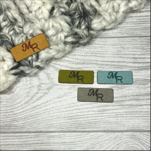 Peut inclure: Plusieurs étiquettes en cuir de différentes couleurs, dont beige, vert olive, bleu sarcelle et gris, chacune estampillée des lettres "MR". Les étiquettes rectangulaires reposent sur une surface en bois gris clair, avec un tissu tricoté en arrière-plan.