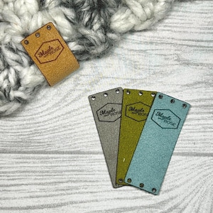 May include: A collection of leather and felt tags with the text "Maple and Rose". The tags are in various colors, including tan, gray, olive green, and teal. The tags have holes for sewing and are displayed on a wooden surface.