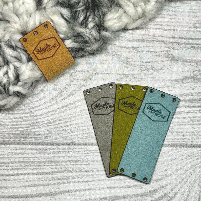 May include: A collection of leather and felt tags with the text "Maple and Rose". The tags are in various colors, including tan, gray, olive green, and teal. The tags have holes for sewing and are displayed on a wooden surface.