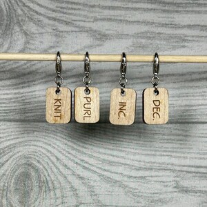 May include: Four wooden knitting stitch markers with silver clasps are displayed on a wooden dowel. Each marker has a rectangular shape with a different knitting term engraved: KNIT, PURL, INC., and DEC.