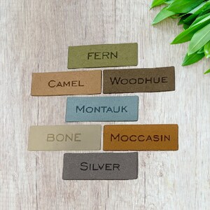 May include: Rectangular leather tags in various colors, including green, tan, and gray, are arranged on a light wood surface. Each tag has a color name embossed on it, such as "FERN," "CAMEL," and "SILVER." Green leaves are in the background.