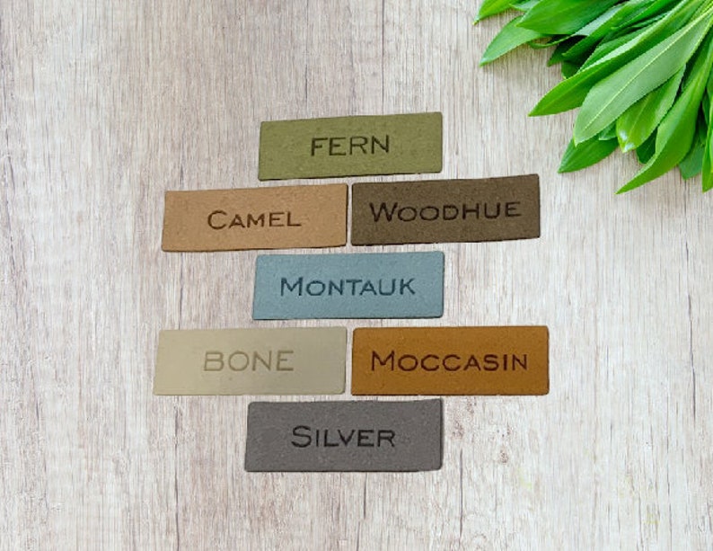 May include: Rectangular leather tags in various colors, including green, tan, and gray, are arranged on a light wood surface. Each tag has a color name embossed on it, such as "FERN," "CAMEL," and "SILVER." Green leaves are in the background.