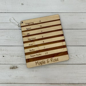 May include: A wooden gauge with a metal ring for hanging. The gauge has a list of yarn weights, including fingering, sport, DK, worsted, aran, bulky, and super bulky, with corresponding needle sizes. The gauge is labelled "Maple & Rose."