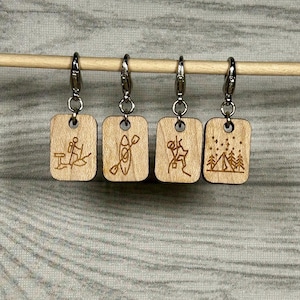 Peut inclure: Quatre breloques rectangulaires en bois avec des fermoirs argentés. Chaque breloque présente une activité de plein air gravée différente : une personne en kayak, une personne sur un paddleboard, une personne en randonnée et un camping avec des arbres.