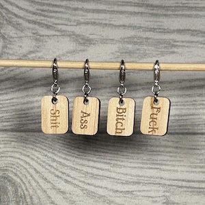 May include: Four wooden stitch markers with silver clasps. Each marker has a rectangular wooden tag with a different word etched into it: "Shit", "Ass", "Bitch", and "Fuck". The markers are hanging on a wooden knitting needle.