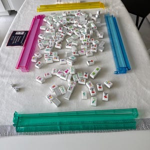 K&ouml;nnte beinhalten: Ein Mahjong-Set mit wei&szlig;en Spielsteinen mit farbenfrohen Symbolen und Zeichen. Das Set enth&auml;lt vier farbige Gestelle in Gelb, Rosa, Blau und Gr&uuml;n sowie W&uuml;rfel und ein Regelbuch. Das Spiel befindet sich auf einer wei&szlig;en Tischdecke.