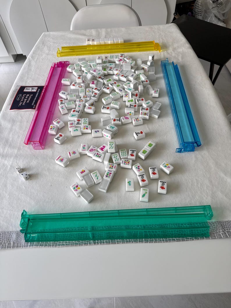 K&ouml;nnte beinhalten: Ein Mahjong-Set mit wei&szlig;en Spielsteinen mit farbenfrohen Symbolen und Zeichen. Das Set enth&auml;lt vier farbige Gestelle in Gelb, Rosa, Blau und Gr&uuml;n sowie W&uuml;rfel und ein Regelbuch. Das Spiel befindet sich auf einer wei&szlig;en Tischdecke.