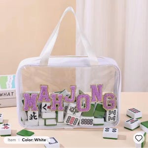 K&ouml;nnte beinhalten: Eine wei&szlig;e, transparente Tragetasche mit dem Wort "MAHJONG" in lila, glitzernden Buchstaben. Die Tasche enth&auml;lt Mahjong-Steine in Gr&uuml;n, Wei&szlig; und Rot. Die Tasche hat wei&szlig;e Griffe.