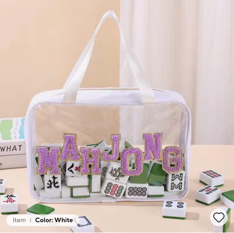K&ouml;nnte beinhalten: Eine wei&szlig;e, transparente Tragetasche mit dem Wort "MAHJONG" in lila, glitzernden Buchstaben. Die Tasche enth&auml;lt Mahjong-Steine in Gr&uuml;n, Wei&szlig; und Rot. Die Tasche hat wei&szlig;e Griffe.