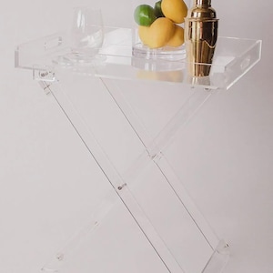 May include: A clear acrylic folding side table with a rectangular top and a cross-legged base. The table holds a clear glass, a glass jar with lemons and limes, and a gold cocktail shaker. The table is designed for serving drinks and snacks.
