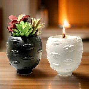 Pode incluir: Dois porta-velas decorativos, um preto e outro branco, com um padrão em relevo de lábios. O porta-velas branco tem uma vela acesa dentro. Uma pequena planta suculenta está no porta-velas preto.