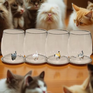 May include: Four clear glass cups with a saucer base, each containing a small figurine of a cat. The cats are in various poses and colors, including white, gray, and yellow. The cups are arranged in a row on a wooden surface, with several cats looking at the cups.