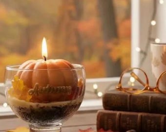 Może przedstawiać: Płonąca świeca w kształcie dyni znajduje się w przezroczystej szklanej misce, z wytłoczonym słowem Autumn na boku. Obok leżą dwie ułożone w stos stare książki, bursztynowe okulary do czytania oraz ceramiczny kubek z motywem liści.