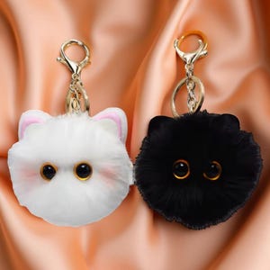 May include: Two fluffy cat keyrings, one white with pink ears and one black. Both have large, round, black eyes with gold pupils.