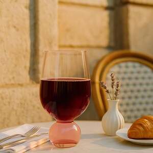 May include: A wine glass with a pink base and a clear bowl, filled with red wine. The glass sits on a white marble table with a gold rim, next to a small white vase with lavender and a plate with a croissant. A folded white napkin and silverware are also on the table.