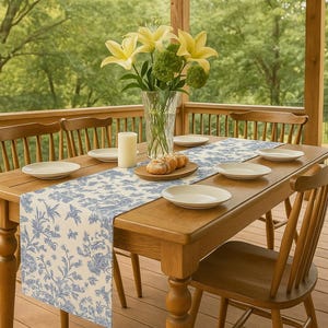 Könnte beinhalten: Ein Holztisch, der für eine Mahlzeit auf einer Veranda gedeckt ist. Der Tisch hat einen blau-weißen Blumenschmuck, weiße Teller, eine Kerze und eine Vase mit gelben Blumen. Holzstühle umgeben den Tisch, und der Hintergrund zeigt eine grüne Landschaft.