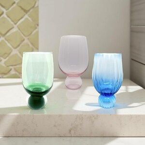 May include: Three colorful stemware glasses. One is green, one is pink, and the third is blue. The green glass has a dark green base. The pink glass has a pink base. The blue glass has a blue base and vertical ridges.