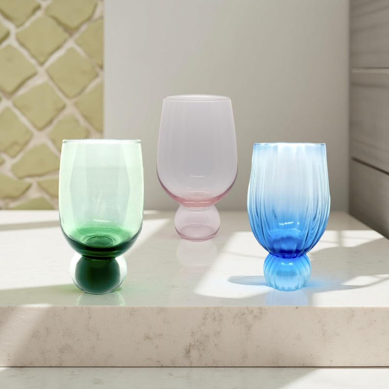 May include: Three colorful stemware glasses. One is green, one is pink, and the third is blue. The green glass has a dark green base. The pink glass has a pink base. The blue glass has a blue base and vertical ridges.