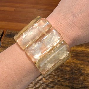 May include: A stretch bracelet composed of three large, rectangular, translucent amber-colored glass beads. The beads feature a smooth, polished surface with internal cloudy inclusions that catch the light, creating a warm, golden glow.