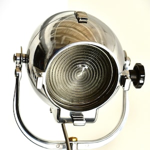 May include: A chrome-plated studio spotlight with a large, round reflector and a circular, ribbed lens. The spotlight is mounted on a tall, adjustable stand.